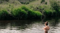 a woman standing in the middle of a body of water next to a lush green hillside