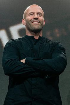 a bald man with his arms crossed standing in front of a screen and smiling at the camera