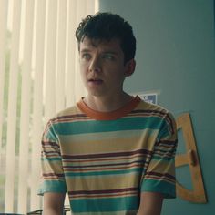 a young man standing in front of a window wearing a striped shirt and looking at the camera