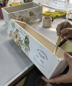 a person holding a box with flowers painted on it and scissors in their hand next to the box