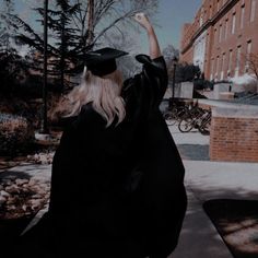 a woman in a black graduation gown and cap is sitting on the sidewalk with her hand up