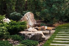 a table and chairs in a garden with rocks, grass and trees behind it is a stone path that leads up to an outdoor seating area