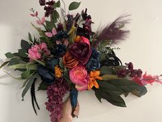 a hand holding a bouquet of flowers on a wall with feathers and foliage around it