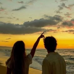 a man and woman standing on top of a beach next to the ocean at sunset