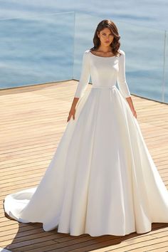 a woman in a white wedding dress standing on a wooden deck near the ocean with her hands on her hips