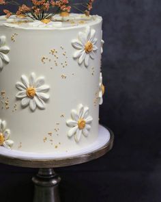 a white and gold decorated cake with flowers on it's side, sitting on a metal stand