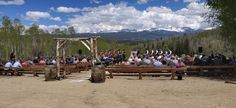 a group of people sitting on wooden benches in the middle of a dirt field with mountains in the background