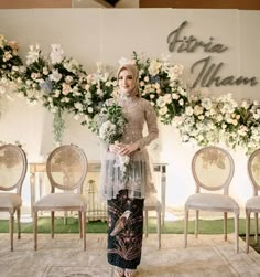 a woman standing in front of chairs with flowers on the wall behind her, holding a bouquet