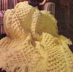 a cat sitting on top of a couch under a yellow crocheted blanket next to a chair