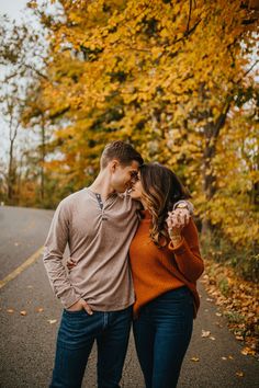 an engaged couple standing on the road in front of trees with fall foliage behind them