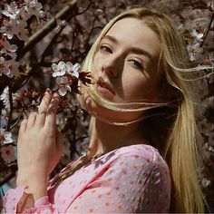 a woman with long blonde hair standing in front of a tree filled with pink flowers