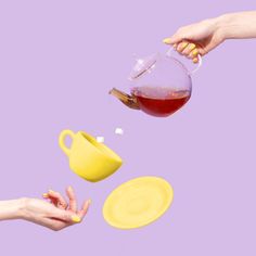 two hands reaching for a pitcher of liquid from a yellow cup and saucer on a purple background