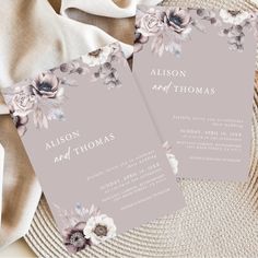 two wedding cards with flowers on them sitting on top of a white cloth covered table