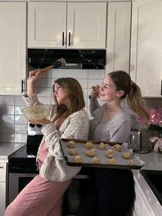 two women in the kitchen with cookies on trays and one holding a cookie bar