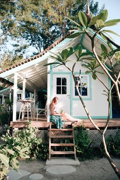 a woman sitting on a porch in front of a small white house with blue trim