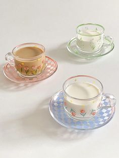 three glass cups and saucers with flowers on them, one is filled with milk