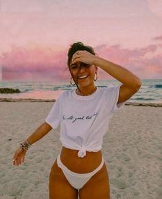 a woman standing on top of a sandy beach next to the ocean with her hands behind her head