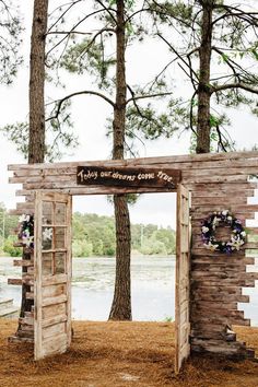 a wooden structure with flowers on the front and side is surrounded by pine trees, along with a lake in the background