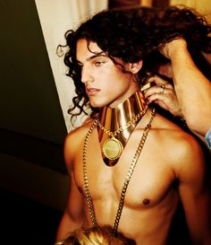 a shirtless man is getting his gold collared necklace put on by another person