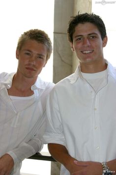 two young men standing next to each other in front of a building and one is wearing a white shirt