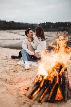 a man and woman sitting next to a campfire on the beach with their arms around each other