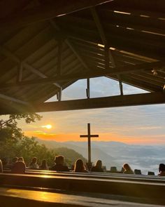 people sitting in pews watching the sun rise over mountains and valleys with a cross on top