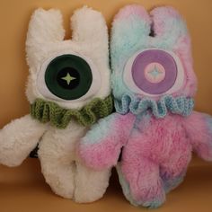 two stuffed animals are sitting next to each other on a brown background, one is wearing a green bow tie and the other has a pink teddy bear with an eye