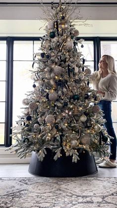 a woman standing next to a white christmas tree in a living room with black and silver ornaments