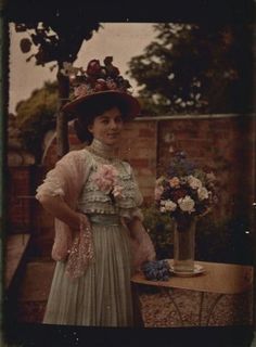 an old photo of a woman in a dress and hat standing next to a table with flowers