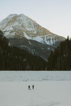 two people are walking through the snow in front of a mountain