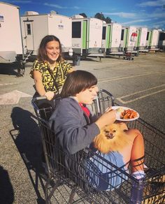 two people sitting in a shopping cart with a small dog on the front and one person holding a plate