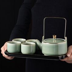 a person holding a tray with four cups and a tea kettle on it, in front of a black background