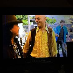 two men standing in front of a tv screen with one man wearing a hat and the other smiling