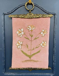 a pink wall hanging with gold trim and flowers on it's side, in front of a blue door