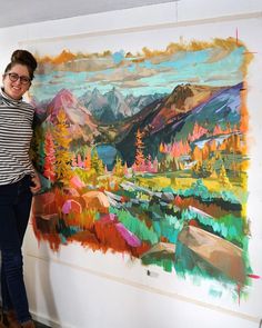 a woman standing in front of a painting with mountains and trees painted on the wall