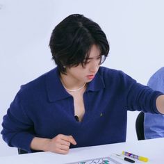 a woman sitting at a table with markers and pens in front of her, while another man looks on