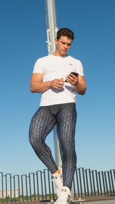 guy wearing a white t-shirt and carbon fibre print leggings and white sneakers holding and looking at his phone on a sunny day while standing up against a metal pole outside Casual High Stretch Tights For Jogging, Casual Moisture-wicking Sports Tights, Moisture-wicking Casual Sports Tights, Casual Moisture-wicking Tights For Gym, Casual Tight Gym Tights, Casual Nylon Training Leggings, Casual Nylon Leggings For Sports, Casual Nylon Leggings For Training, Casual Tight Tights For Gym