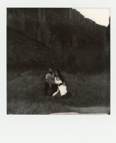 an old photo of two people hugging in the grass near a mountain side with trees