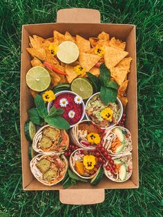 an open box filled with tacos, salsa and chips on top of green grass