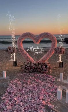 a heart - shaped arrangement on the beach with candles and flowers in front of it