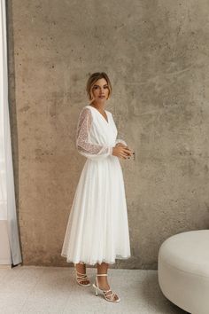 a woman standing in front of a wall wearing a white dress with sheer sleeves and heels