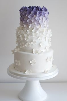 a three tiered white cake with purple flowers on the top and bottom, sitting on a pedestal