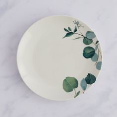 a white plate with green leaves painted on the front and side, sitting on a marble surface