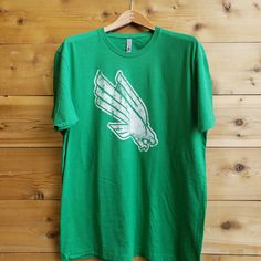 From the heartland to the coasts, wear your Mean Green on your sleeve with your new favorite North Texas t-shirt from us at Nudge Printing! Every shirt is printed by hand with eco-friendly inks on ethically and sustainably sourced garments and then inspected and packaged individually so you know you're getting the best every time. Our shirts are 60% cotton, 40% polyester, so it feels better, breathes easier, and will last longer than your average 100% cotton t-shirt. It is also preshrunk to supp Green Short Sleeve Team Spirit T-shirt, Green Graphic Print T-shirt For Fan Gear, Casual Green T-shirt For Fan Gear, Green Fan Apparel T-shirt With Screen Print, Green Graphic Tee With Team Name, Green Team Spirit Short Sleeve T-shirt, Green Short Sleeve T-shirt With Team Spirit Style, Green Short Sleeve Top For Team Spirit, Diving Eagle