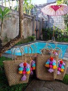 four woven bags with colorful tassels near a swimming pool in a tropical setting