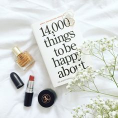 an open book sitting on top of a white sheet next to lipstick and perfume bottles