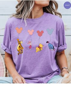 Embark on a whimsical adventure with our Pooh & Gang Balloon Escapade Tee! This charming cotton t-shirt features Winnie the Pooh and his friends soaring skyward in delightful Mickey-shaped balloons. It's a playful nod to classic Disney magic that's perfect for any Pooh fan. Get ready to add a touch of joy and nostalgia to your wardrobe with this fun and colorful tee! Themed Cartoon Print Tops For Disney Trips, Whimsical Cotton Short Sleeve Tops, Fun Crew Neck Tops For Disney Trips, Playful Purple Tops With Cartoon Print, Casual Long Sleeve T-shirt For Disney Trips, Pooh Shirt, Pooh Disney Shirt, Winnie The Pooh Tshirts, Summer Disney Cotton T-shirt