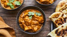 three bowls filled with curry next to pita bread on top of a wooden table