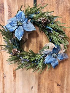 a wreath with blue flowers and pine cones hanging on a wooden wall, ready to be used as a christmas decoration