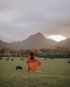 a woman in an orange dress is running through a field with cows and mountains behind her